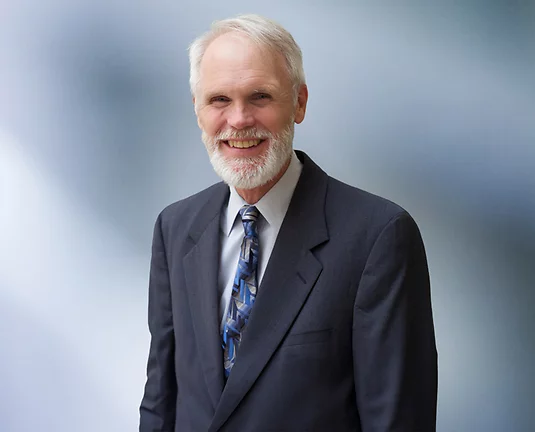 Older man, Chuck Barbieri, with white hair and white beard smiles at camera wearing blue grey blazer, white undershirt and blue and grey patterned necktie.