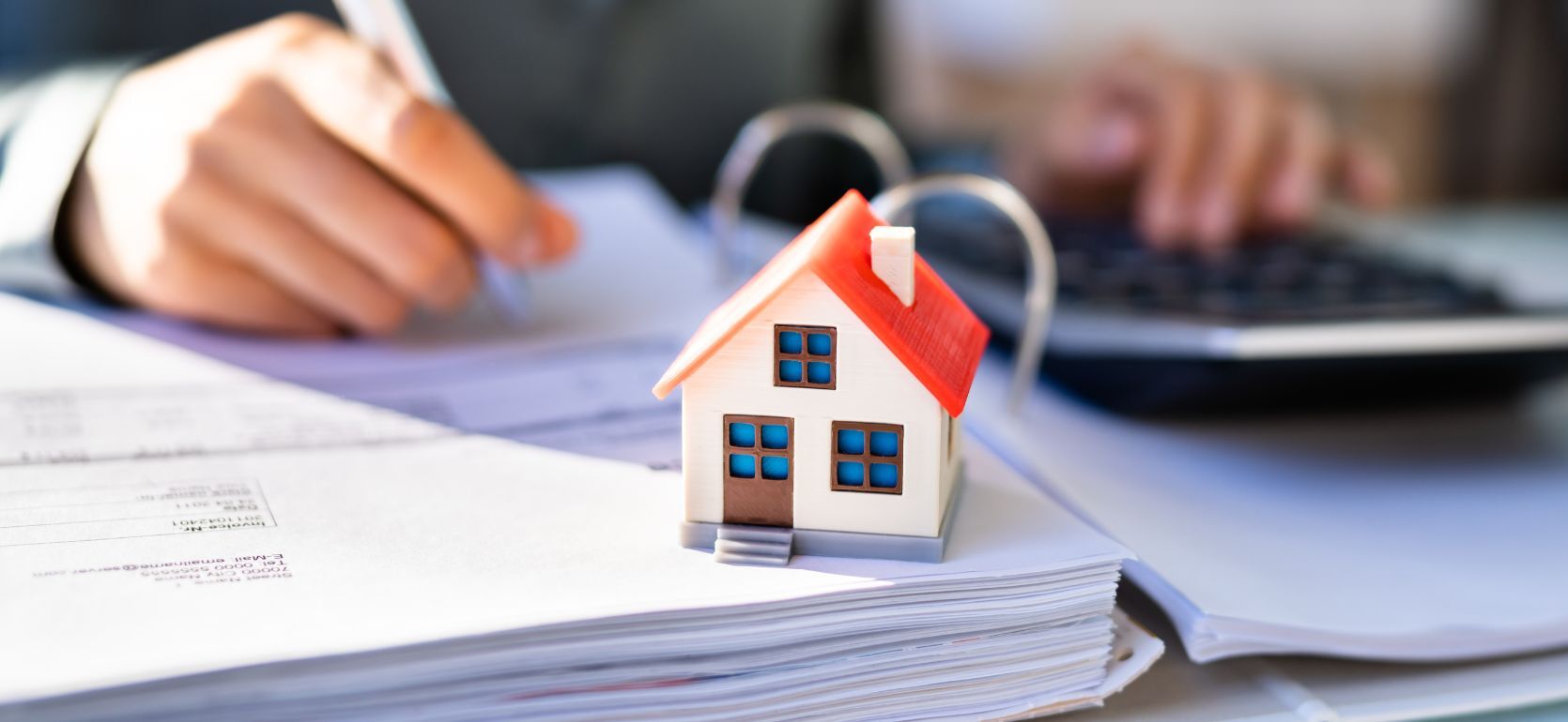 Model house with a red roof sitting on papers while someone writes in the back.