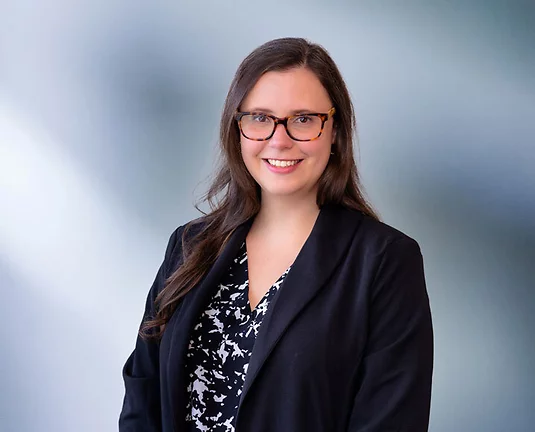 A brown-haired woman, Amy Marinkovski, wearing turtle frame glasses, black sport coat and black and white zigzag pattern shirt, smiles at camera in front of blue gray background