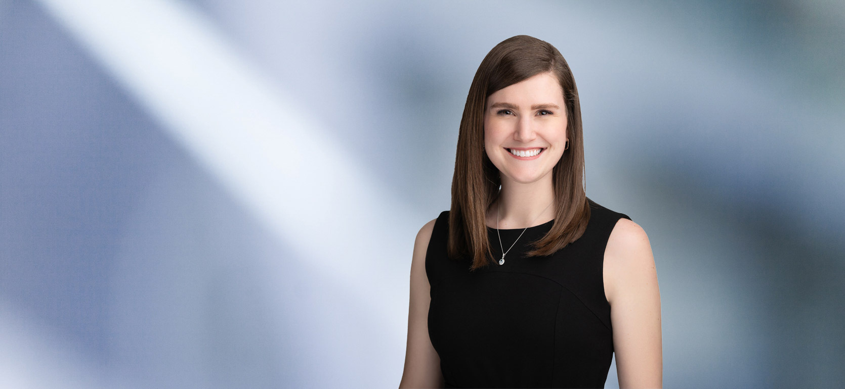 A woman, Amanda Dernovshek, in a sleeveless black dress is smiling at the camera, standing against a blurred blue and white background.