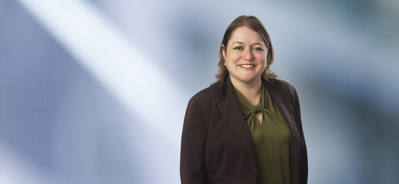 A woman, Anna Gibson, in a dark blazer and green blouse is smiling at the camera, standing against a blurred blue and white background.
