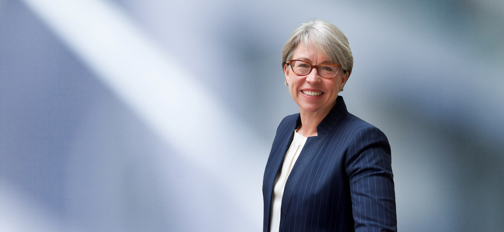 A woman, Deanna Swisher, in a navy pinstripe suit, white blouse, and glasses is smiling at the camera, standing against a blurred blue and white background.