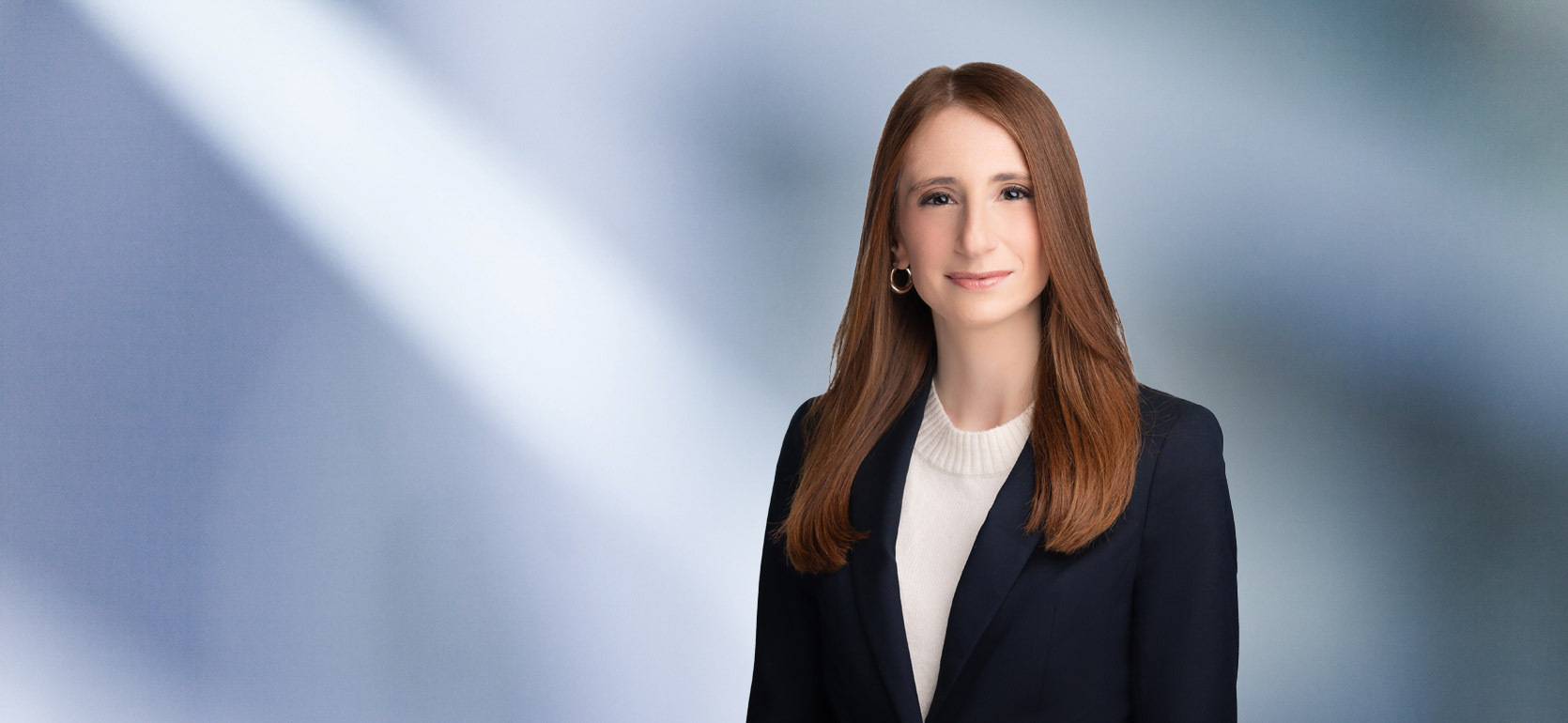 Smiling woman, Dina Kashat, with long, straight auburn hair, wearing a dark blazer over a white top, standing against a soft blue gradient background