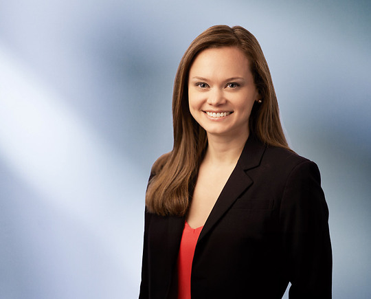 A woman, Leslie Abdoo, in a black blazer and red top is smiling at the camera, standing against a blurred blue and white background.