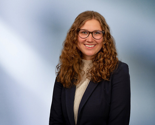 A woman, Mackenzie Almassian, with long, wavy blonde hair, wearing a navy blazer and white top, is smiling at the camera, standing against a blurred blue and white background.”