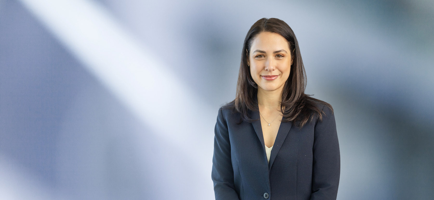 Woman, Alicia Birach, in a dark navy blazer standing against a softly blurred blue and white background, smiling confidently at the camera