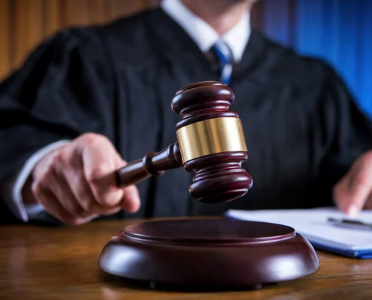 A judge in a courtroom holding a gavel over a wooden sound block, with a legal document and pen on the desk.