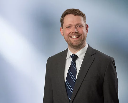 Man with beard, Jon Austin, smiles at camera wearing a gray sport coat and grey striped tie in front of blue gray background