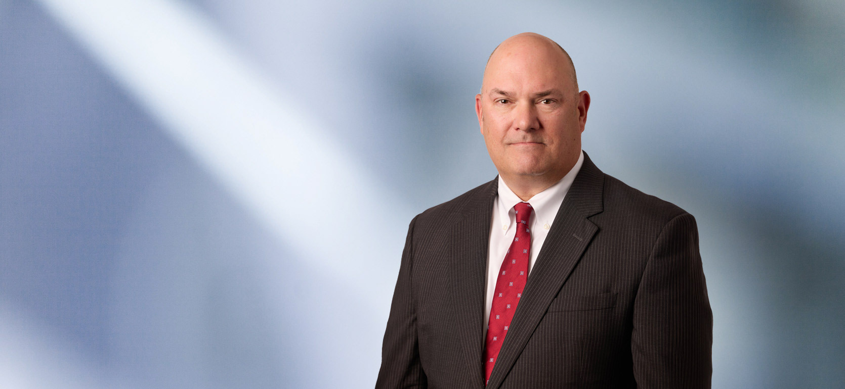 A man, Andrew Vredenburg, in a pinstripe suit, white shirt, and red patterned tie is looking at the camera, standing against a blurred blue and white background.
