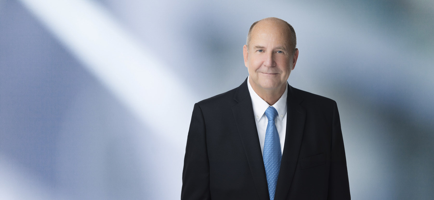 A man, Bryan Cermak, in a dark suit, white shirt, and light blue tie is smiling at the camera, standing against a blurred blue and white background.