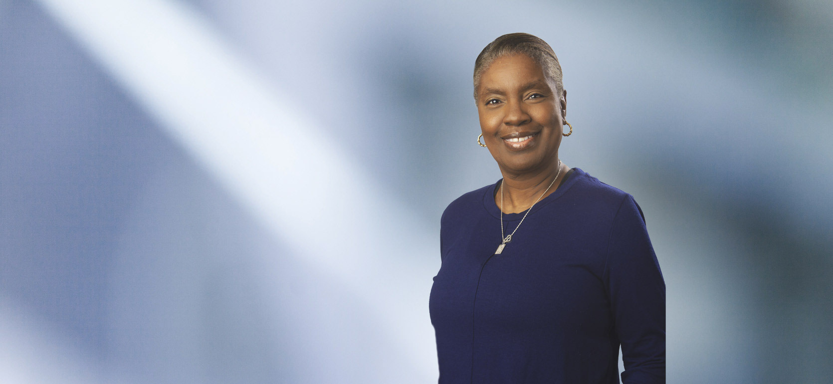 A woman, Gillian Maynard, in a dark blazer and white blouse is smiling at the camera, standing against a blurred blue and white background.