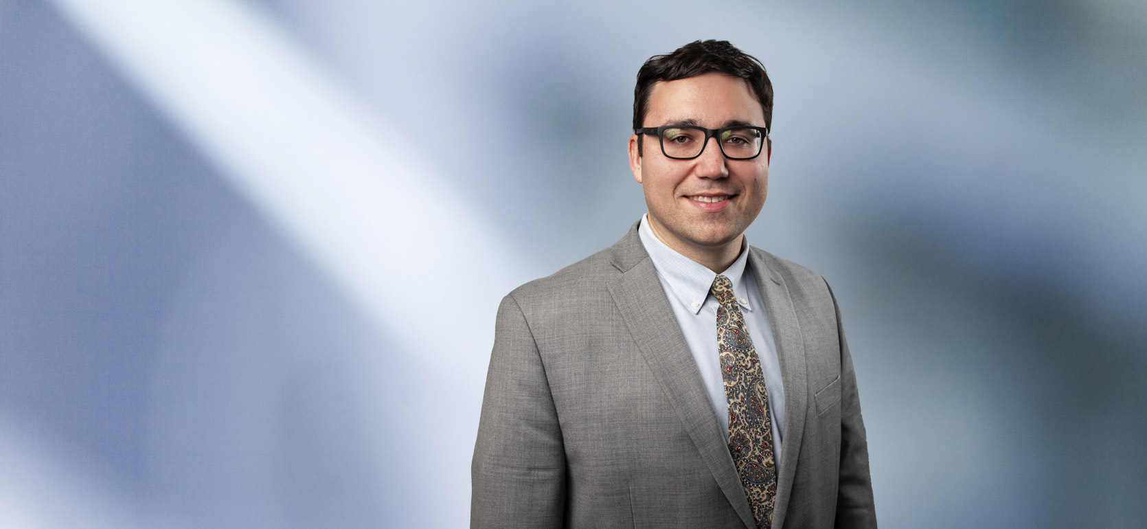 A man, Jeffrey Schultz, in a light gray suit, pale blue shirt, and paisley tie is smiling at the camera, standing against a blurred blue and white background.