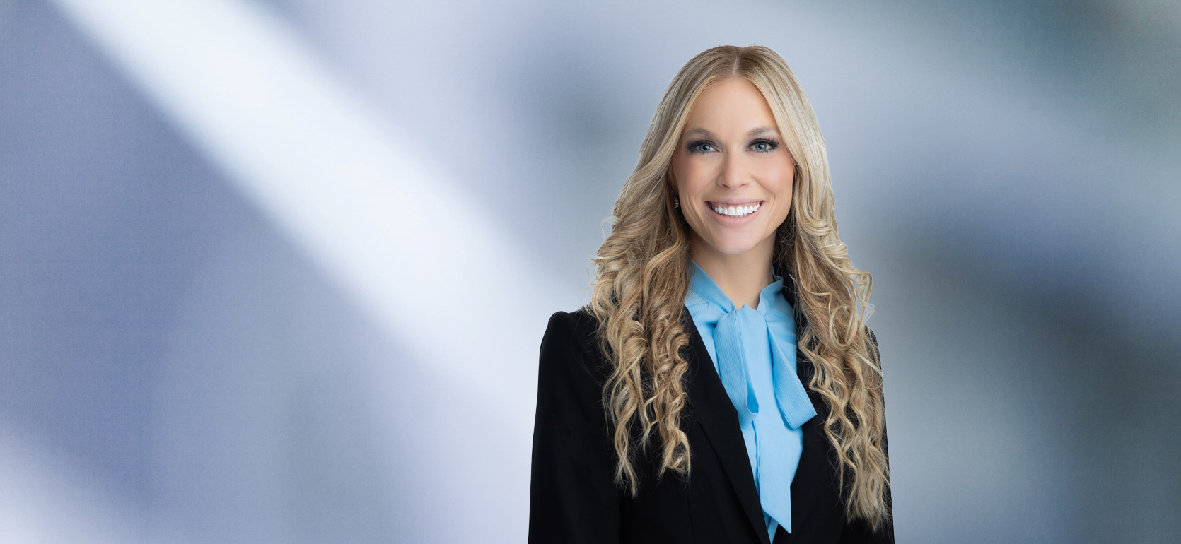 A woman, Jennifer Montasir, in a black blazer and bright blue blouse with long wavy blonde hair is smiling at the camera, standing against a blurred blue and white background.
