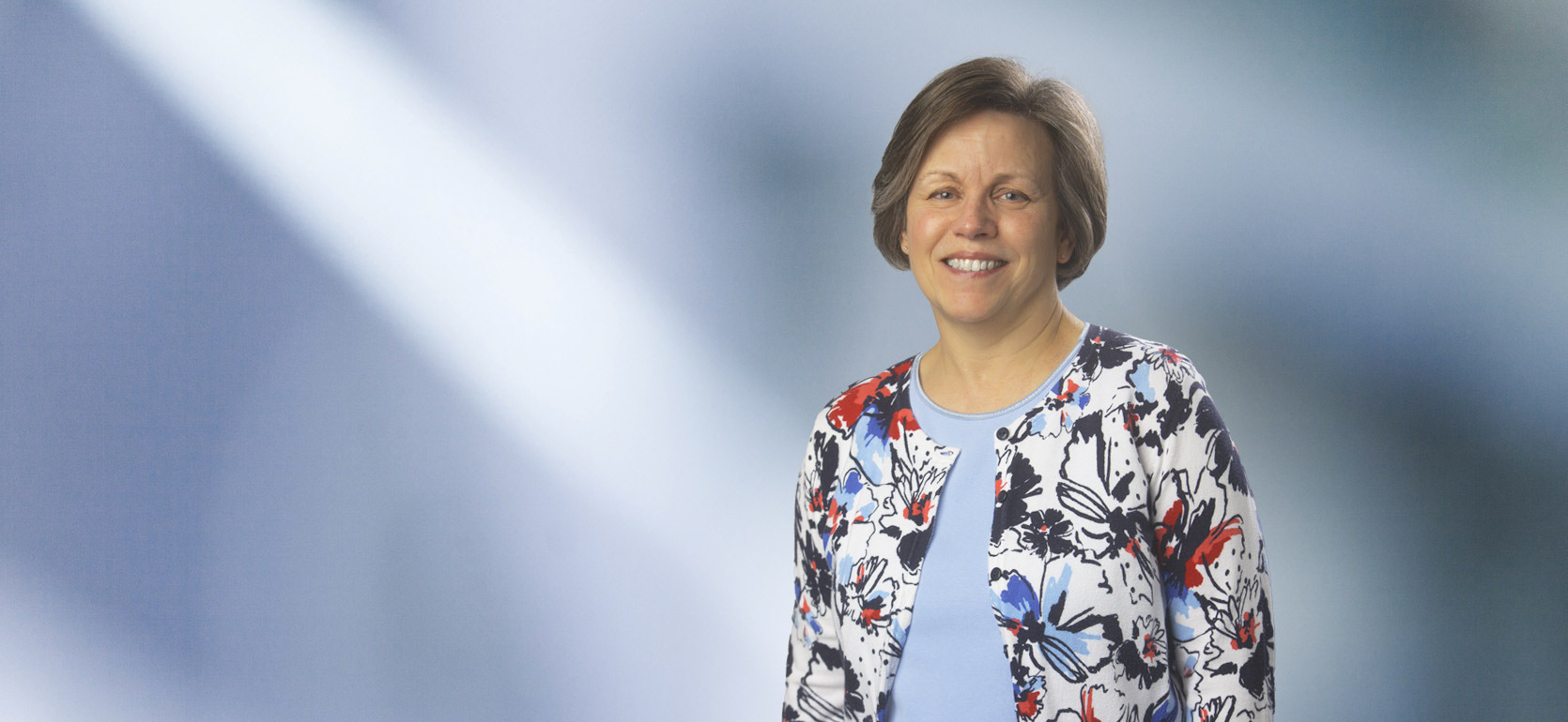A woman, Lori Rosier, with straight, blonde hair, wearing a black blazer and white top, is smiling at the camera, standing against a blurred blue and white background.”