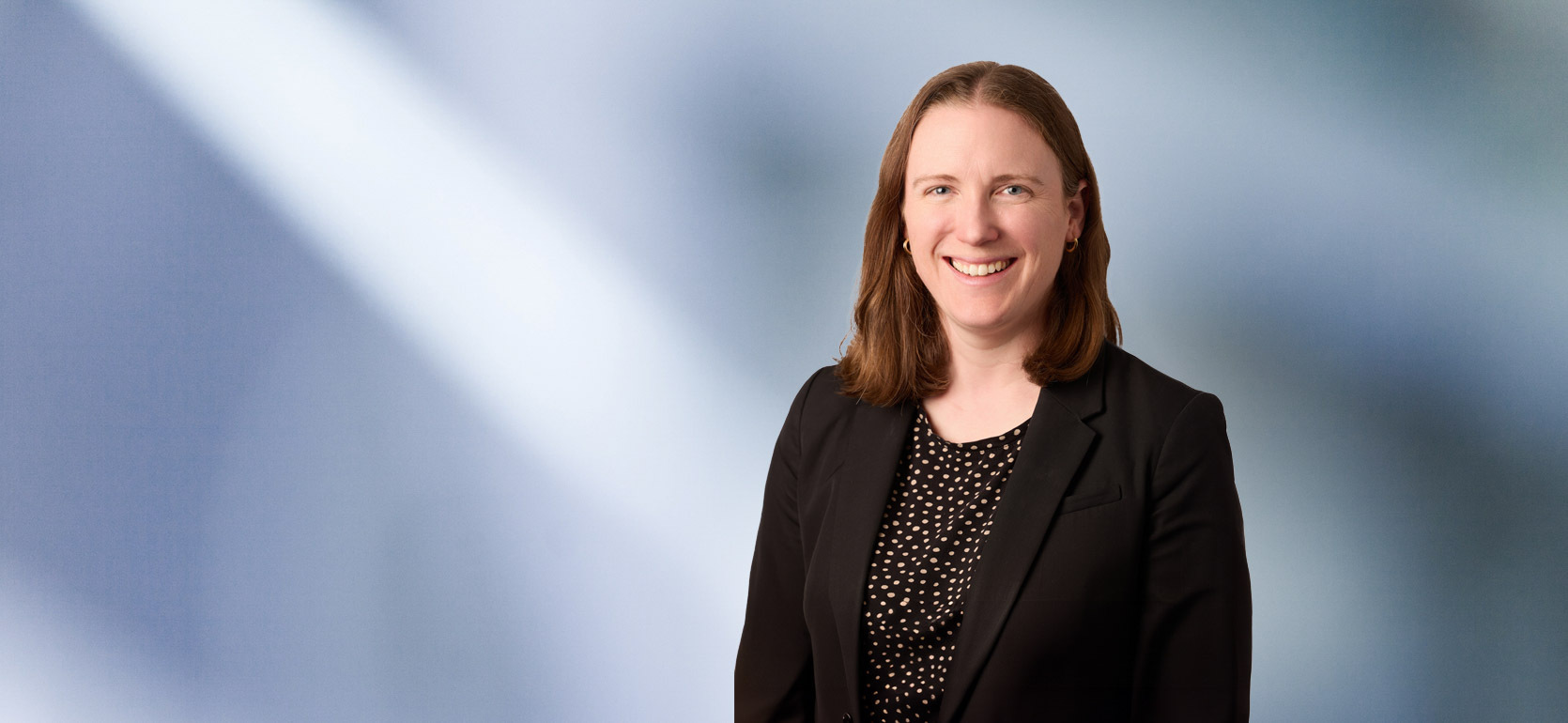 woman, Rachael Kuleima Klein, with brown hair, wearing a black blazer, smiling against a blurred blue and white background"