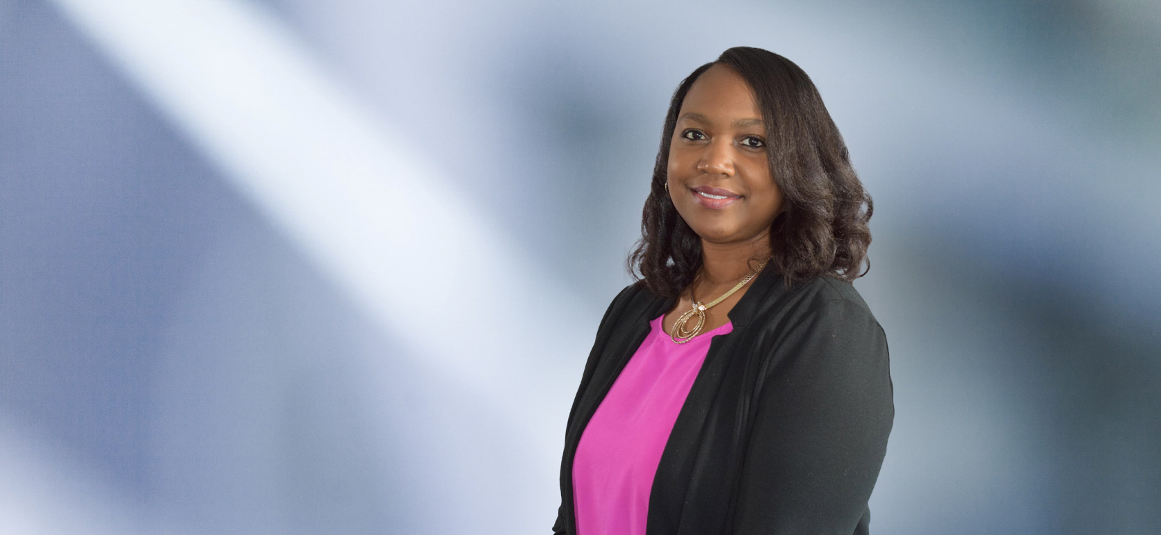 woman, Rekela Wilson, with dark brown hair, wearing a black blazer, smiling against a blurred blue and white background"