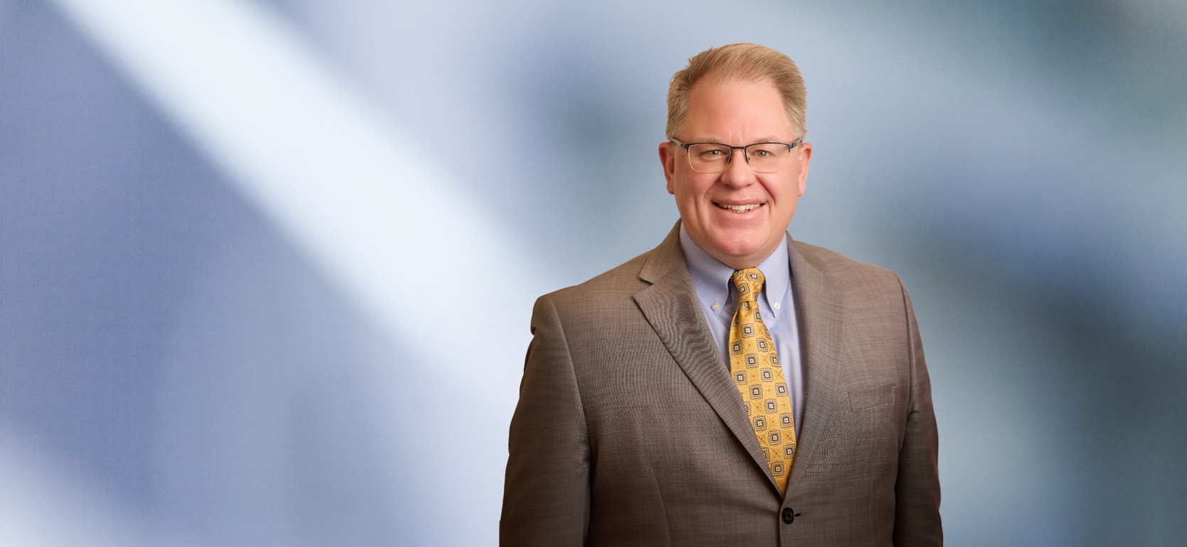 Headshot of a man, Ryan Lamb, with light brown hair, wearing a grey suit and white shirt, smiling against a blurred blue and white background"
