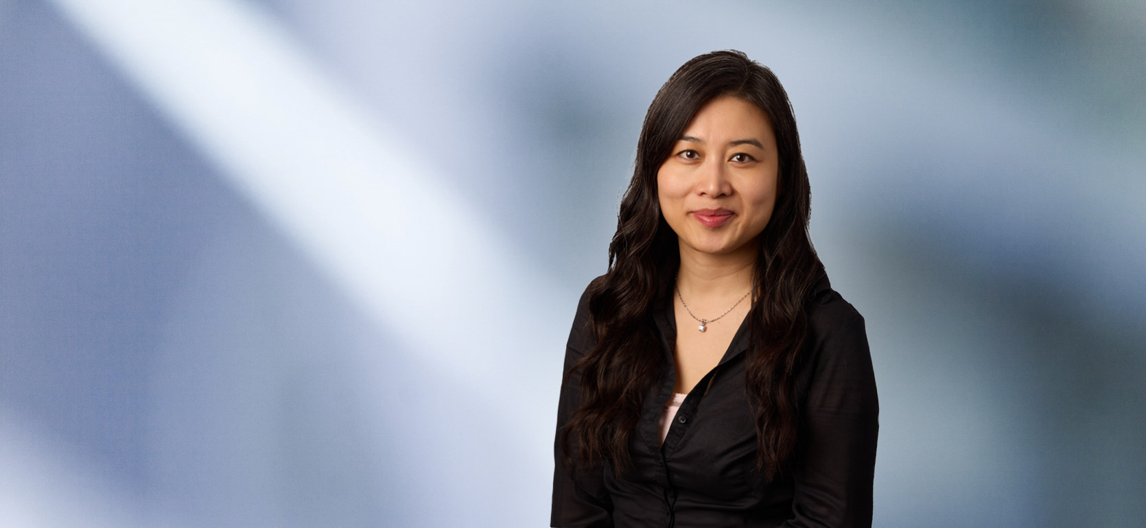 Portrait of a woman, Thailan Hang, with long black hair in a dark blazer, with a blurred blue and white background.