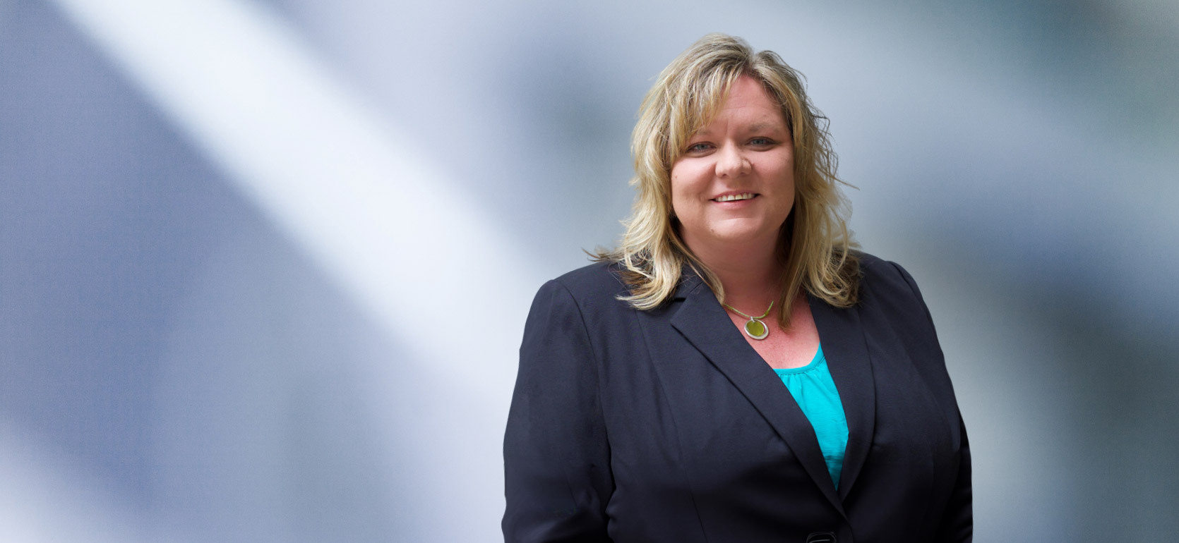 Portrait of a woman, Terry Bolyard, with long blonde hair in a dark blazer, with a blurred blue and white background.