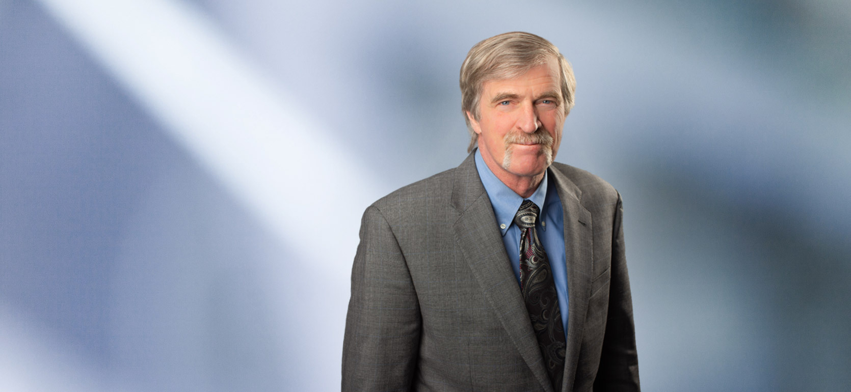 Portrait of a man, Steven Tjapkes, with short light gray hair in a dark blazer, with a blurred blue and white background.