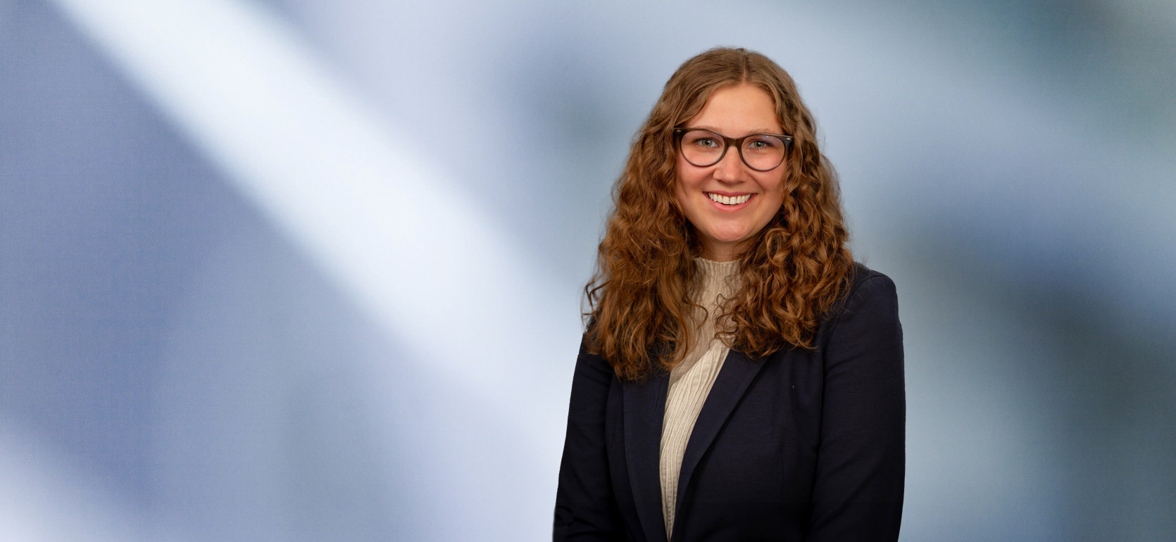 A woman, Mackenzie Almassian, with long, wavy blonde hair, wearing a navy blazer and white top, is smiling at the camera, standing against a blurred blue and white background.”
