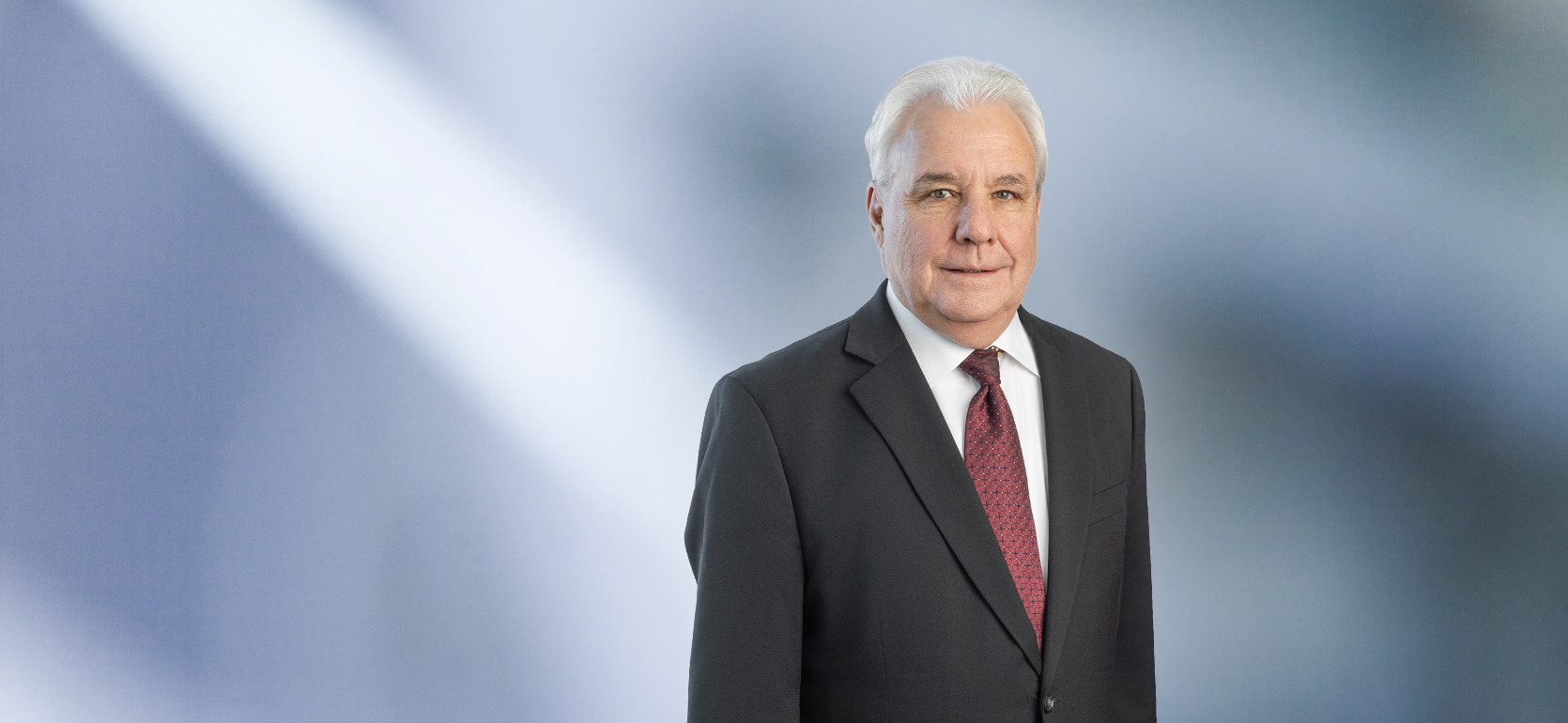 man with white hair wearing a dark suit, white shirt, and maroon patterned tie, standing against a softly blurred blue and white background