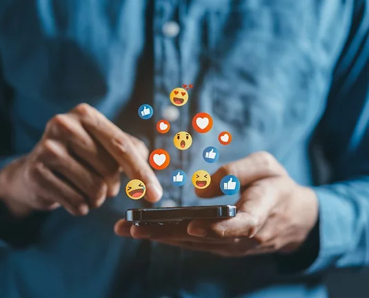 A person wearing a denim shirt uses a smartphone. Floating above the phone are various colorful social media reaction emojis, including hearts, thumbs up, laughing faces, and shocked faces, representing online engagement and interaction.