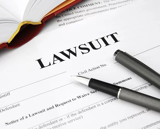 Close-up of a lawsuit document with a pen and an open book on a desk, focusing on the word “LAWSUIT” in bold at the top of the page.