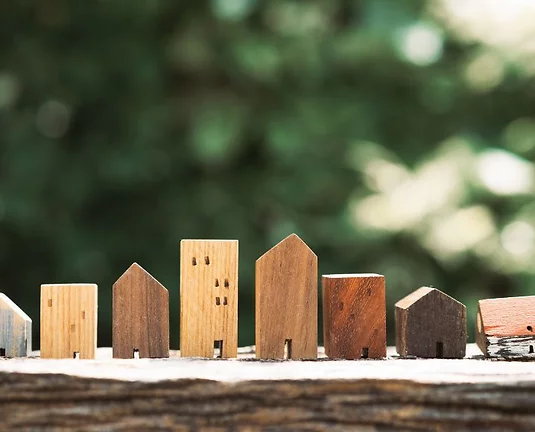 Row of small wooden houses in various shapes and shades, lined up on a rustic wooden surface with a blurred green natural background.