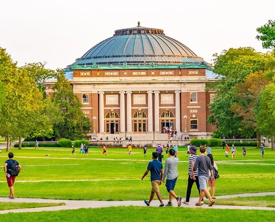 College students walking across a green campus lawn toward a large, domed academic building with classical columns and arched windows, surrounded by trees.