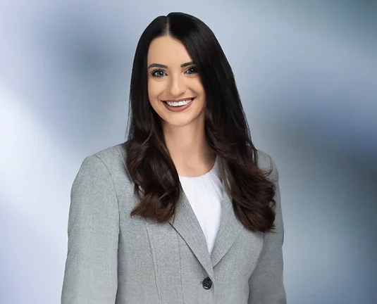 Young women, Melanie Assad, long flowing black hair smiles at camera wearing grey blazer with white blouse.
