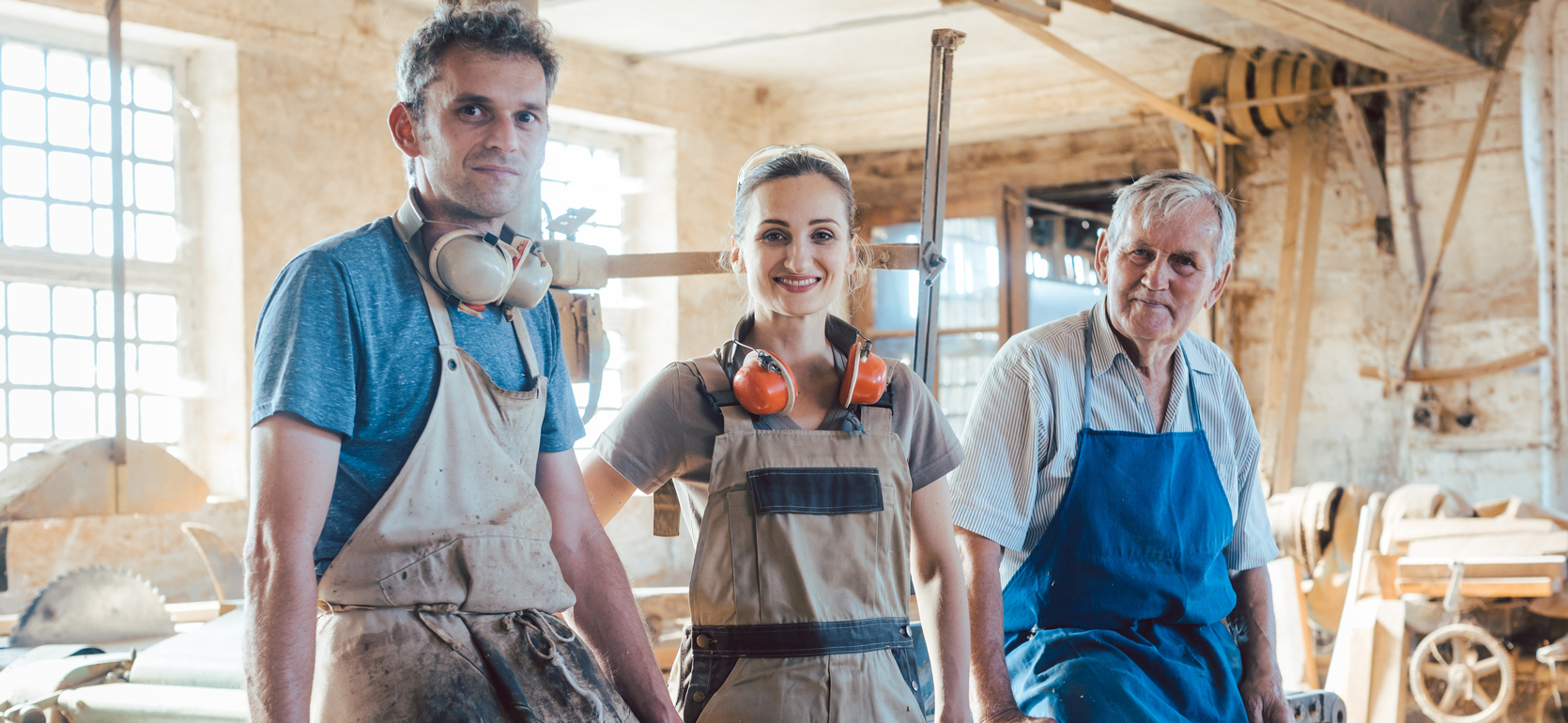 Multiple age generations of a family-owned carpentry business stand and smile at camera wearing aprons in a woodshop surrounded by tools.