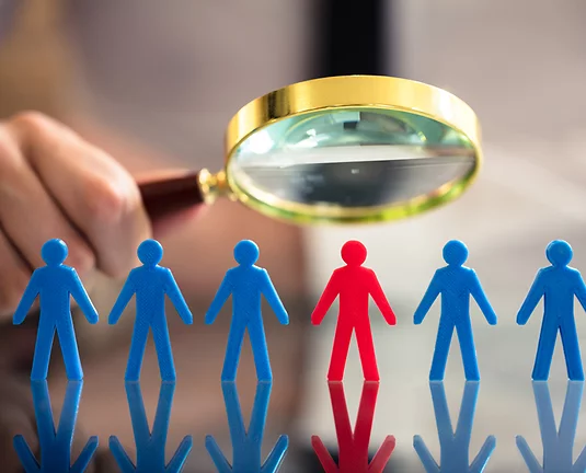 Hand holds magnifying glass over paper cutout figure colored blue with one red one that the glass hovers over. Diversity concept.