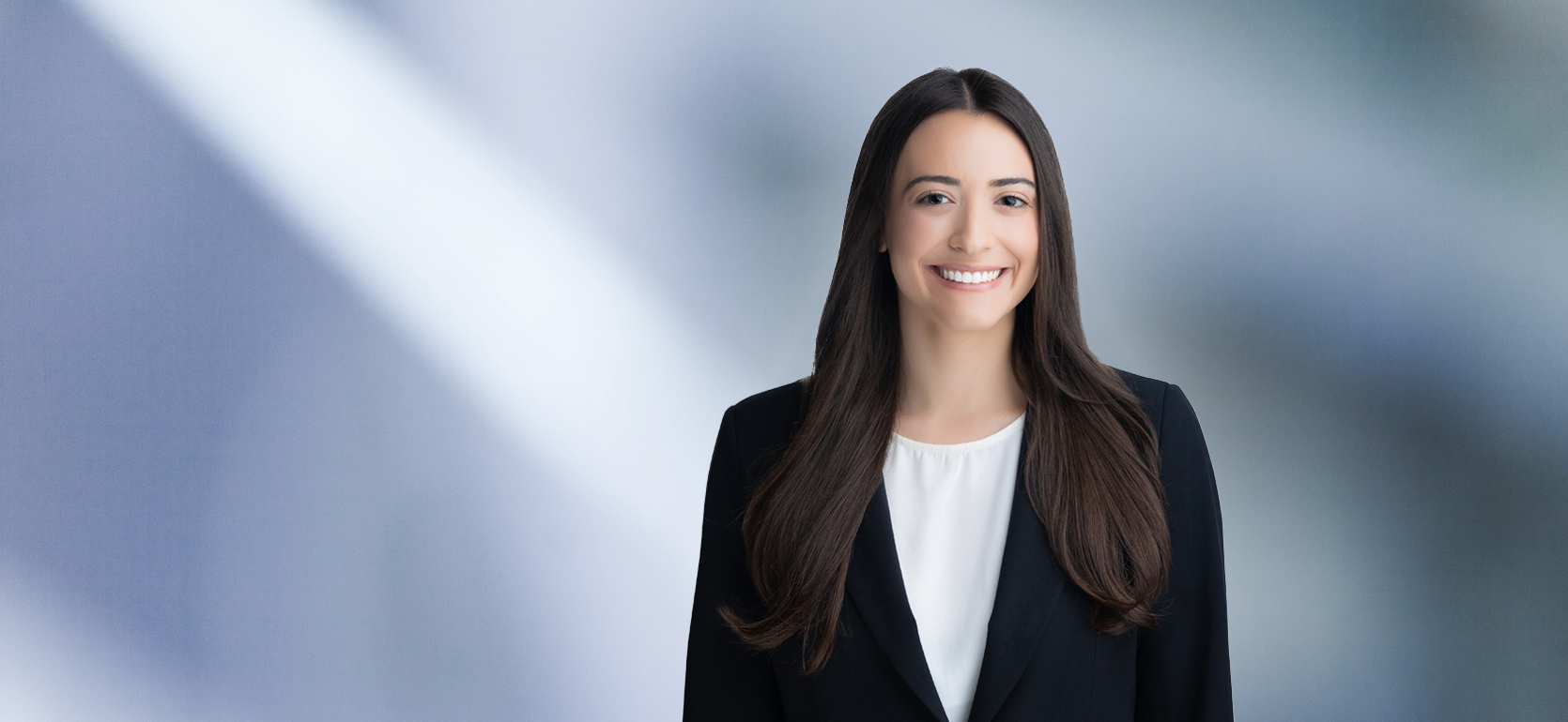 A woman, Danielle Romano, in a dark suit and white blouse is smiling at the camera, standing against a blurred blue and white background.