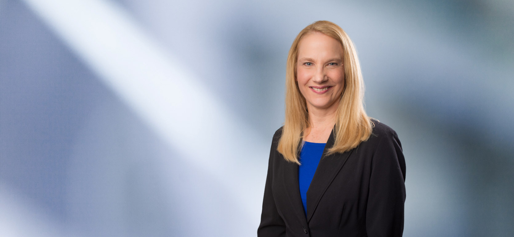 A woman, Lisa Myers, with long blonde hair, wearing a black blazer and bright blue top, is smiling at the camera, standing against a blurred blue and white background.