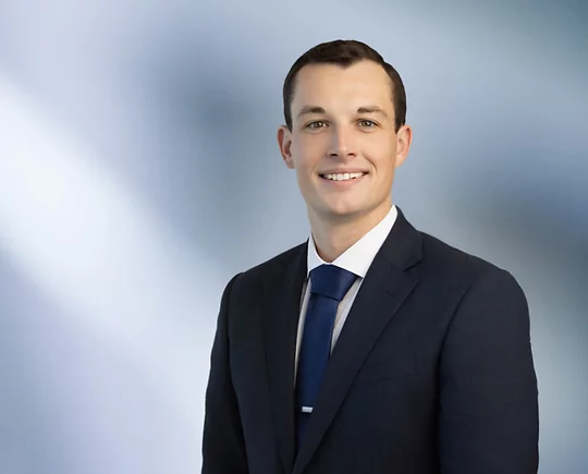A man James Ryan, in a dark suit, white shirt, and blue tie is smiling at the camera, standing against a blurred blue and white background.