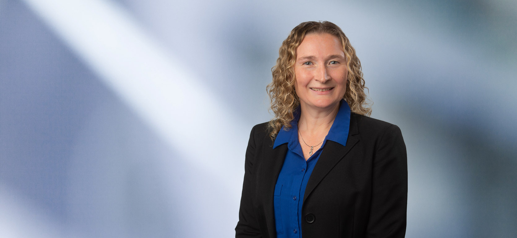 Woman with curly blonde shoulder length hair, Jennifer Knox, wearing black blazer and dark blue collared button-down shirt, smiles at camera in front of blue gray background
