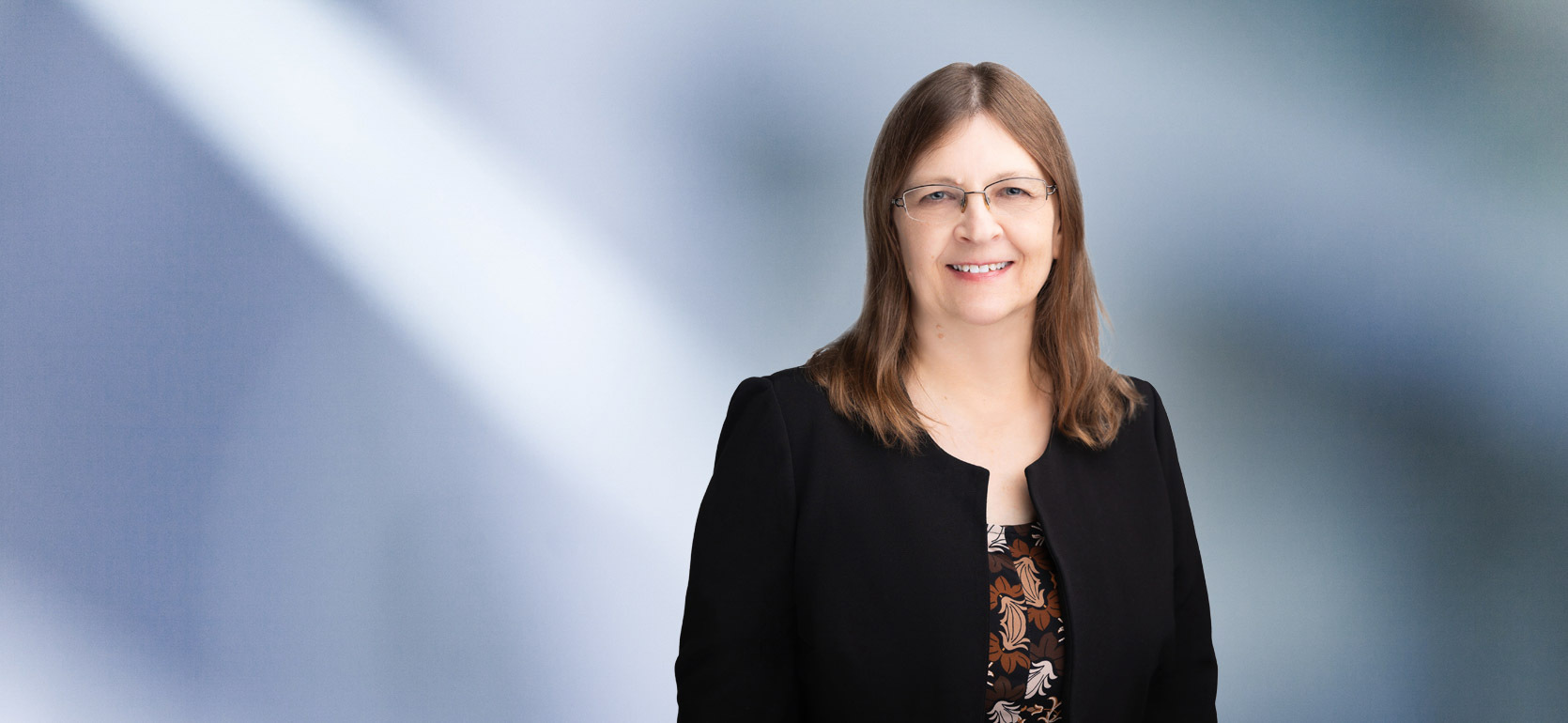 A woman, Beverly Swan, in a black blazer and patterned top is smiling at the camera, standing against a blurred blue and white background.