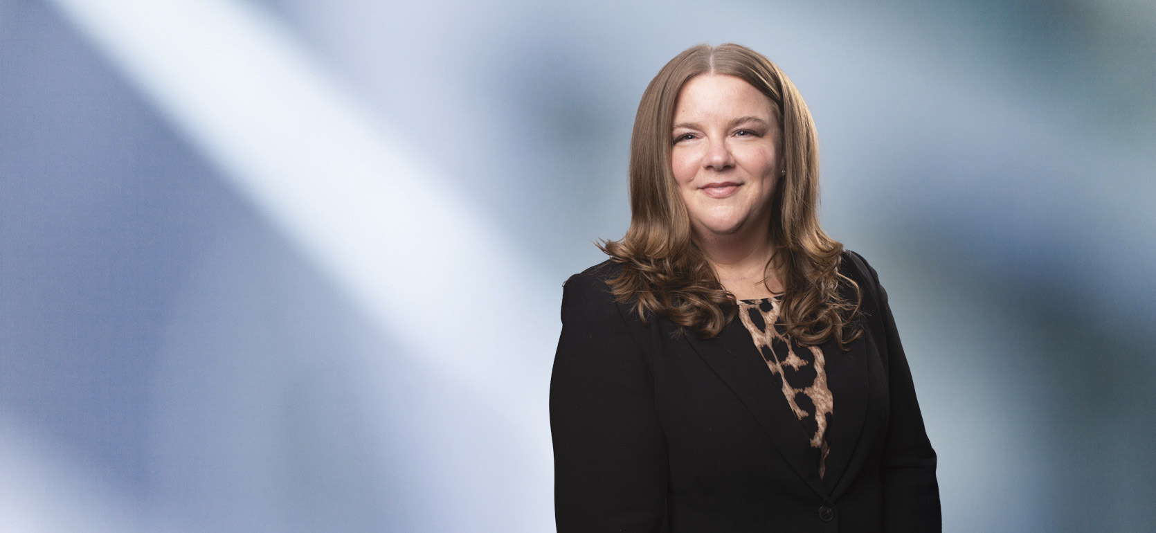 A woman, Cassandra Bowe, in a black blazer and leopard print top is smiling at the camera, standing against a blurred blue and white background.