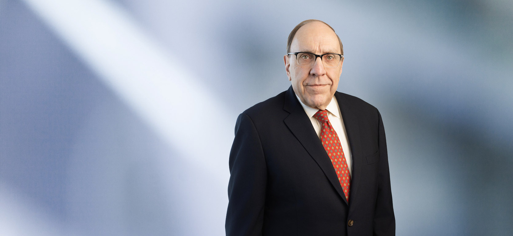 man, Brian Renaud, wearing a dark suit, white shirt, red patterned tie, and glasses, standing against a softly blurred blue and white background, looking confidently at the camera