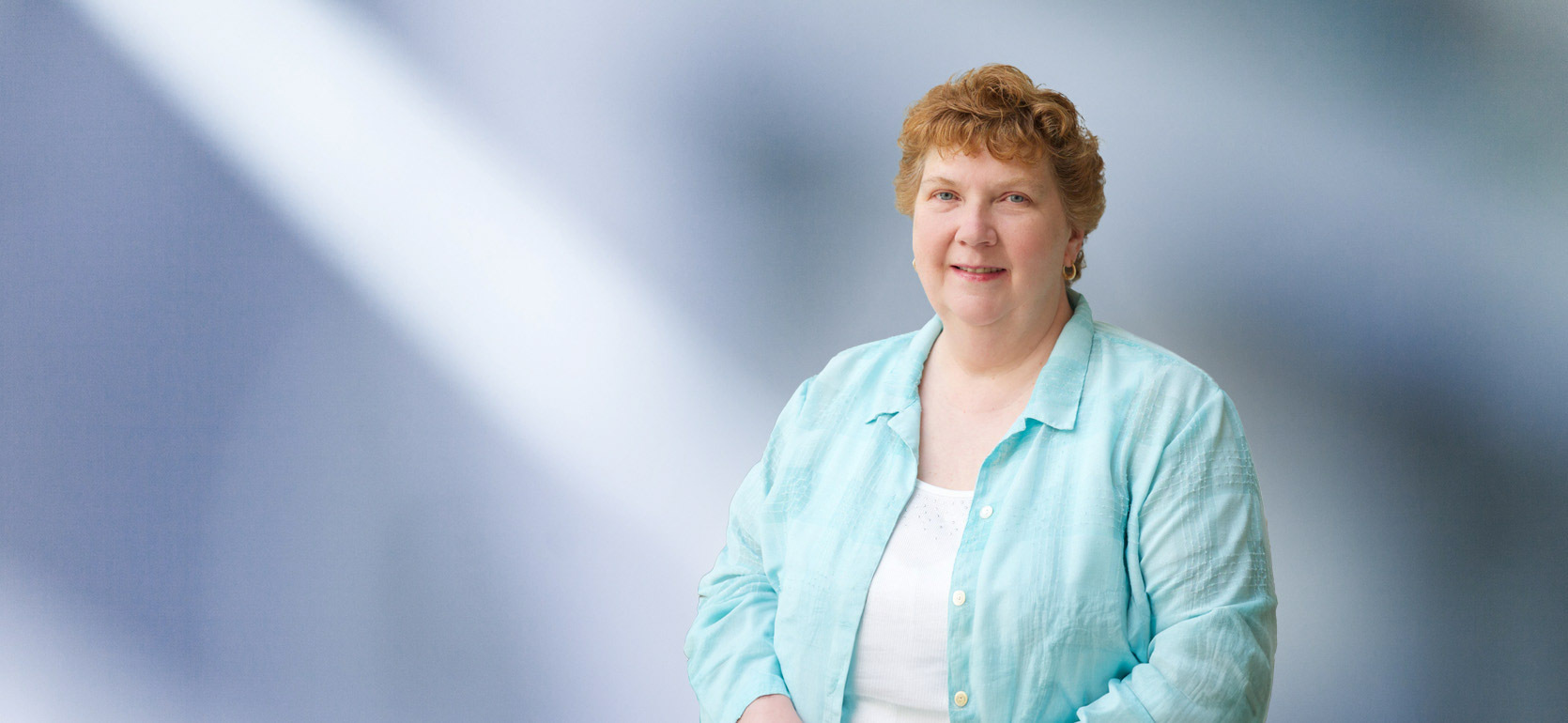woman, Carole Schneider, wearing a light turquoise blouse, standing against a softly blurred blue and white background, smiling warmly at the camera.
