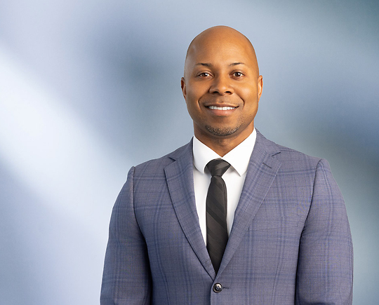 A bald African American man, Ray Littleton, wearing a blue tweed pattern sport coat with a white undershirt and black striped tie is smiling at the camera, standing against a blurred blue and white background.