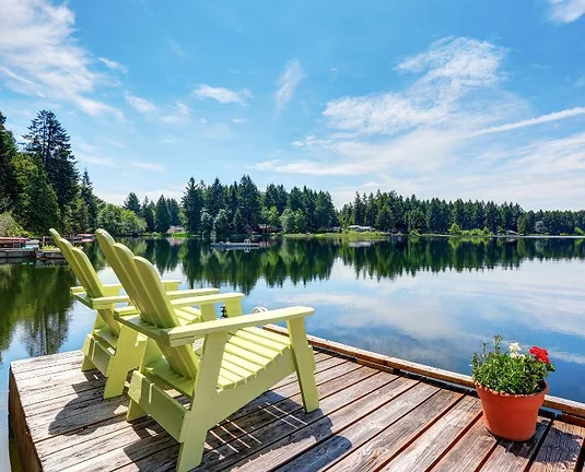 Two yellow chairs sitting on a wood dock overlooking a lake with large green trees on the shore.