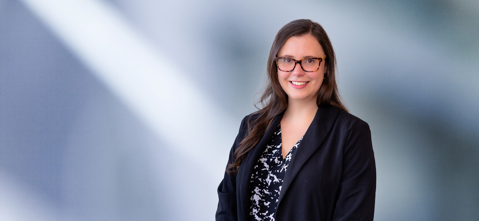 A brown-haired woman, Amy Marinkovski, wearing turtle frame glasses, black sport coat and black and white zigzag pattern shirt, smiles at camera in front of blue gray background