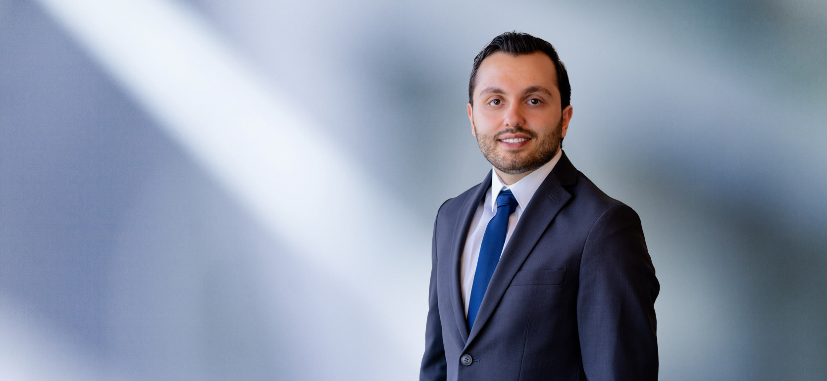 A young man, Badri Yono, wearing a blue-gray blazer, white shirt and solid blue time, smiles at camera in front of blurred blue-white background