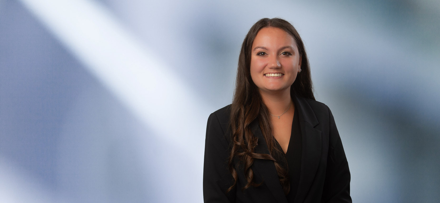 A young dark-haired woman, Rachel Conner, wearing a black sport coat and black blouse, smiles at camera in front of blurred blue-white background