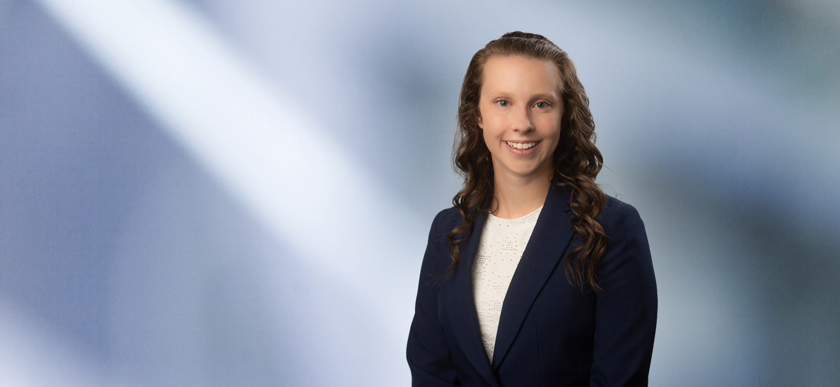 A young woman, Allison Shudark, wearing a black sport coat and white lace blouse, smiles at camera in front of blurred blue-white background