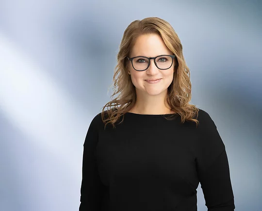 A woman, Megan Cochrane, with shoulder-length, straight blonde hair, wearing a black long-sleeved shirt and black framed glasses, is smiling at the camera, standing against a blurred blue and white background.