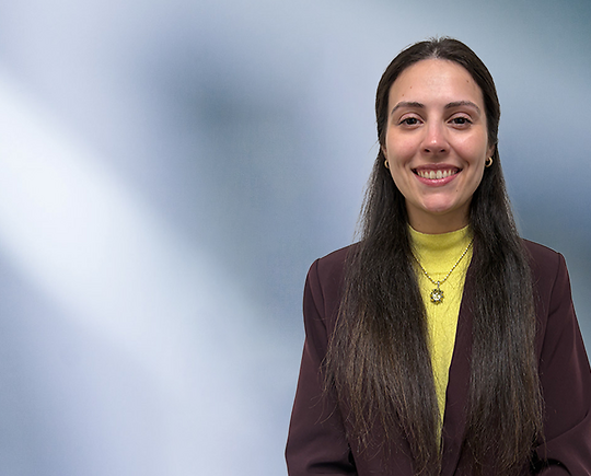 Woman with long brown hair, Stephanie Angelo, wearing a yellow turtleneck, necklace and purple blazer, smiles at camera in front of blurred blue gray background.
