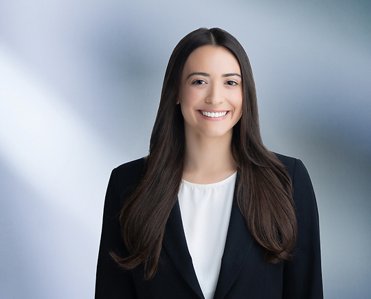 A woman, Danielle Romano, in a dark suit and white blouse is smiling at the camera, standing against a blurred blue and white background.